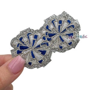 May include: Two silver glitter cheerleading pom poms with blue and white embroidery. The pom poms are attached to each other.