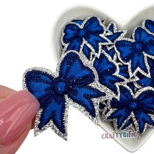 May include: A close-up of a silver glitter bow with a blue center. The bow is surrounded by other similar bows in a heart-shaped bowl.