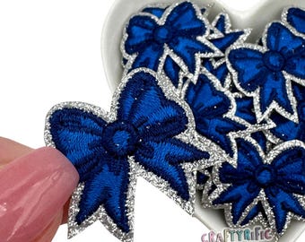 May include: A close-up of a silver glitter bow with a blue center. The bow is surrounded by other similar bows in a heart-shaped bowl.