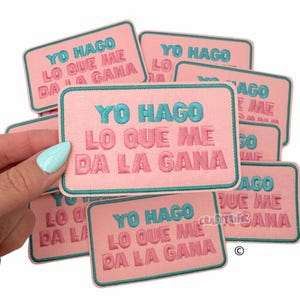May include: A stack of pink and teal iron-on patches with the text "YO HAGO LO QUE ME DA LA GANA" in Spanish. The patches are embroidered with pink and teal thread.