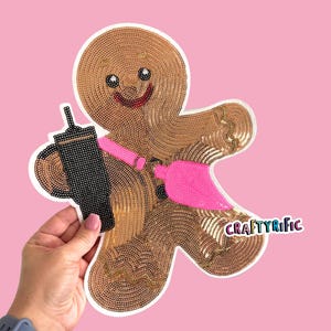 May include: A gingerbread man-shaped applique with a smiling face, holding a black coffee cup and a pink paintbrush. The applique is made of brown and gold sequins, with the text "CRAFTYrific" in the lower right corner.