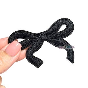 May include: A black embroidered bow patch with a detailed design. The patch is held by a hand with a light pink nail polish. The text "CRAFTYRIFIC" is visible on the right side of the bow.