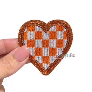 May include: Heart-shaped patch with a checkered orange and white pattern. The patch is outlined with a sparkly, bronze-colored border. The text "CRAFTYRIFIC" is visible on the lower right side of the patch.