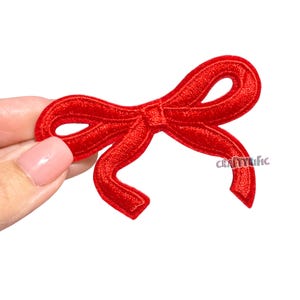 May include: A bright red embroidered bow patch with a detailed design. The patch is in the shape of a bow with two loops and two tails. The text "CRAFTYRIFIC" is visible on the right side of the bow. The patch is held against a white background.