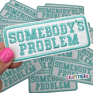 May include: Embroidered patch with the text "SOMEBODY'S PROBLEM" in turquoise lettering on a white background. The patch has a turquoise border and is rectangular in shape. Several patches are visible.