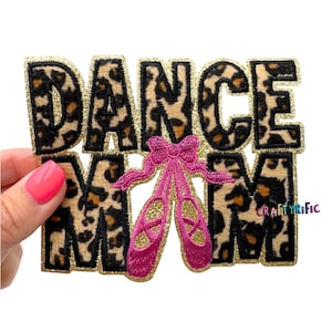 May include: A patch with the words "DANCE MOM" in leopard print letters with gold glitter trim. Pink ballet shoes with gold accents are centered below the word "DANCE". The patch is held by a hand with a pink painted nail.