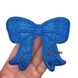 May include: A blue glitter bow applique with a stitched outline. The bow has a classic bow shape with two loops and two streamers. The text "CRAFTYRIFIC" is visible on the right streamer.