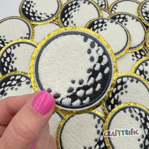 May include: A set of white and black embroidered iron-on patches with a gold glitter trim. The patches are round and feature a design of a golf ball with a half-moon shape.