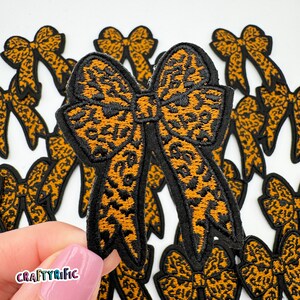 May include: Embroidered bow patches with a leopard print design in shades of black and orange. The patches have a black border and are arranged on a white background. The CraftyRific logo is visible in the lower left corner.