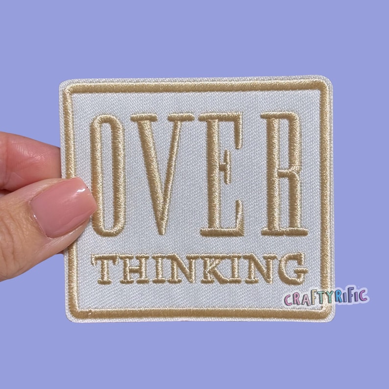Op de afbeelding: Een vierkante, witte patch met goudkleurige geborduurde tekst en rand. De tekst luidt "OVER THINKING" in een vetgedrukte lettertype met schreef. De patch wordt tegen een lichtpaarse achtergrond gehouden. De rechter benedenhoek heeft de tekst "CRAFTYRIFIC".