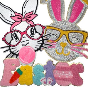 K&ouml;nnte beinhalten: Zwei wei&szlig;e Hasen-Applikationen mit rosa und gelben Akzenten. Ein Hase tr&auml;gt eine rosa Herzbrille und bl&auml;st eine rosa Blase. Der andere Hase tr&auml;gt eine gelbe Brille. Das Wort "Ostern" ist in bunten Buchstaben mit einer rosa Karotte geschrieben.