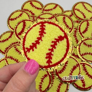 May include: A close-up of a pile of yellow and red felt softball patches with gold glitter trim. The patches are about 2 inches in diameter and have a fuzzy texture.