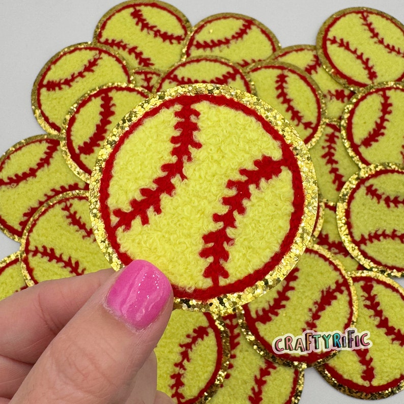 K&ouml;nnte beinhalten: Nahaufnahme eines Stapels gelber und roter Filz-Softball-Patches mit goldfarbenem Glitzerbesatz. Die Patches haben einen Durchmesser von etwa 5 cm und eine flauschige Textur.