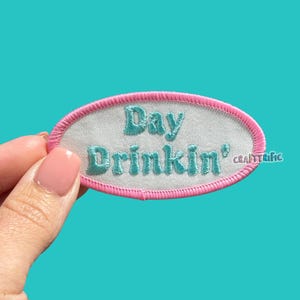 May include: An embroidered patch with the words "Day Drinkin'" in teal lettering. The patch has a white background with a pink oval border. The patch is held by a hand against a teal background.