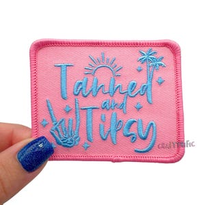 May include: Pink embroidered patch with the words "Tanned and Tipsy" in light blue script. The design includes a sun, palm tree, and a skeleton hand. The patch has a pink border and the text "Crafterific" is in the bottom right corner.