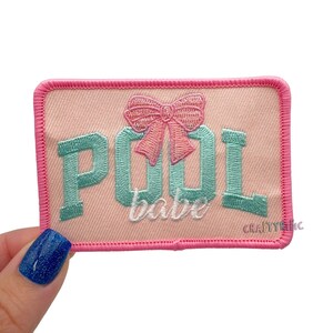 May include: A rectangular patch with a pink background and a pink border. The patch features the words "POOL babe" in teal and white script, with a pink bow above the word "babe". The patch is likely an iron-on or sew-on patch.