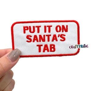 May include: A rectangular white patch with a red border and the embroidered text "PUT IT ON SANTA'S TAB" in red. The patch has rounded corners and the text "CRAFTYRIFIC" in the bottom right corner.