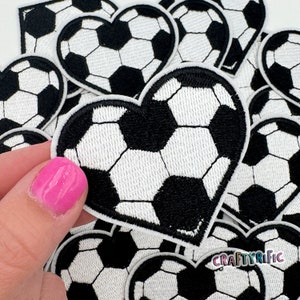 May include: A close-up of a pile of black and white heart-shaped patches with a soccer ball design. The patches are made of felt and have a stitched edge.