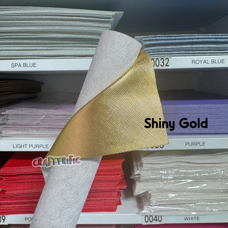 May include: A roll of white and shiny gold fabric is displayed, with the words "Shiny Gold" printed on the gold fabric. The fabric is on a shelf with other colored fabrics, including Spa Blue, Royal Blue, Light Purple, and White.