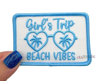 May include: A rectangular patch with a white background and a blue border. The patch features the text "Girl's Trip BEACH VIBES" in blue script, along with heart-shaped sunglasses with palm trees inside.