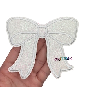 May include: A white glittery bow-shaped patch with a stitched outline. The patch has a central knot and two long, flowing tails. The text "CRAFTYRIFIC" is visible on the right tail. This patch is ideal for adding a decorative touch to clothing or accessories.