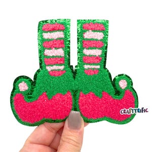 May include: Green and pink felt elf shoes with white stripes. The shoes are decorated with glitter and have a fuzzy texture.