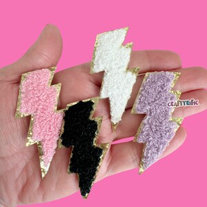 May include: Four fuzzy lightning bolt patches with gold glitter trim. The patches are pink, white, black, and purple.