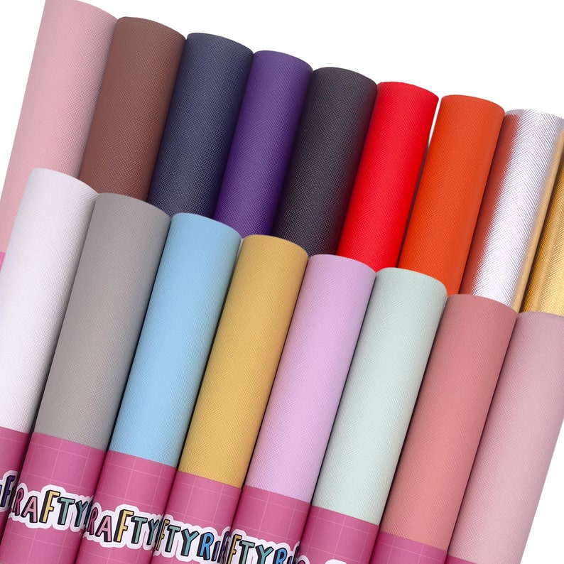 May include: A collection of rolled sheets of textured craft paper in various colors, including pink, brown, navy, purple, red, orange, and gold. The rolls are arranged in a diagonal pattern, with the edges of the paper visible. The bottom of the rolls have a pink label with the text "CRAFTYRIBBON".