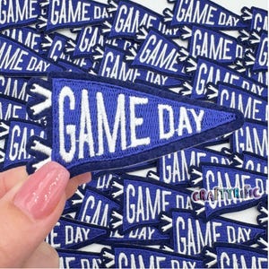 May include: A close-up of a pile of blue felt pennant patches with white embroidery that reads "GAME DAY".