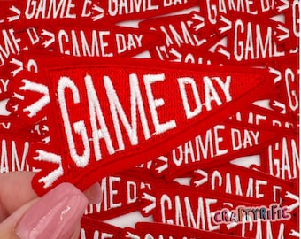 May include: A close-up of a pile of red felt pennants with white embroidered text that reads "GAME DAY".
