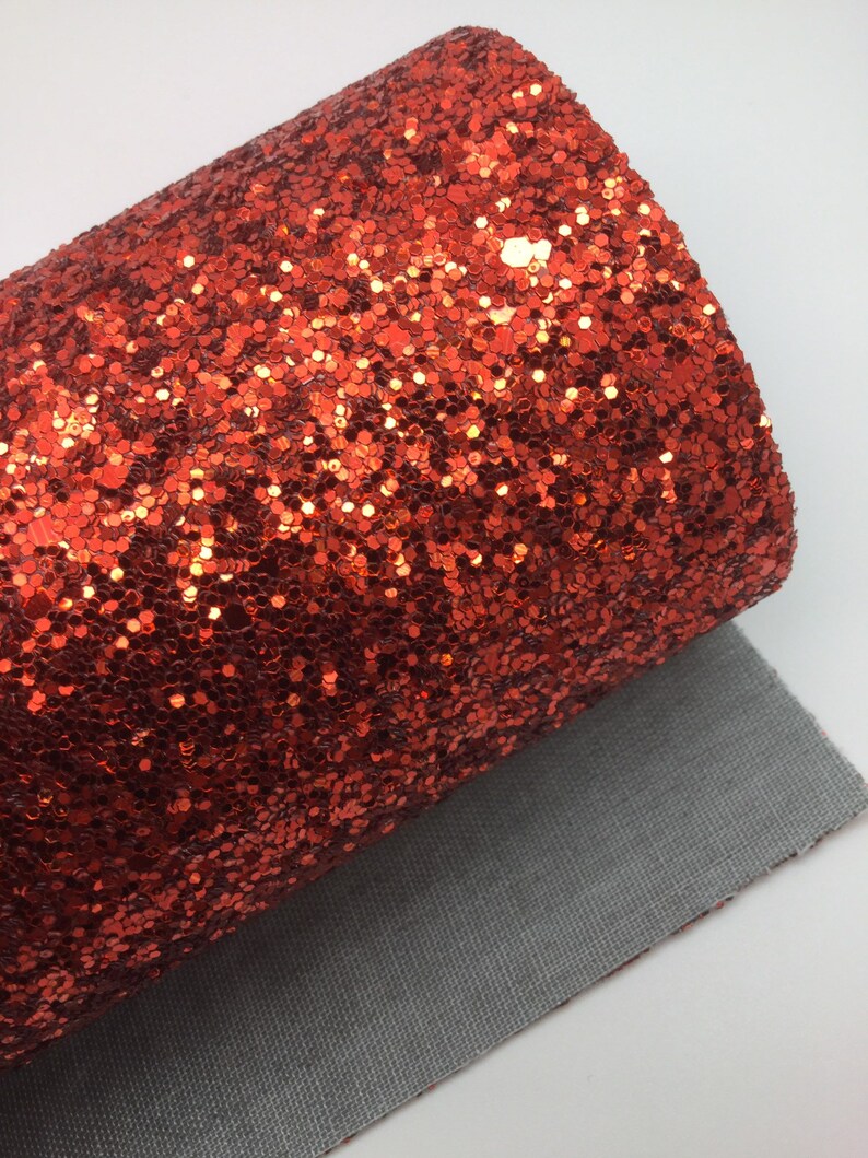Op de afbeelding: Een rol rode glitterstof met een textuur. De stof is gevoerd met een grijs materiaal.