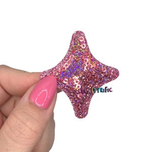 May include: A pink sequined star-shaped embellishment with a four-point design. The star has a glittery texture and is held against a white background. The text "CRAFTYRIFIC" is visible on the star.