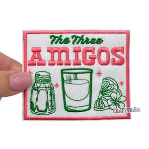 May include: White embroidered patch with pink text that reads "The Three Amigos". The patch features a green salt shaker, a green shot glass, and a green lime wedge.