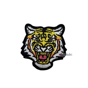 May include: Embroidered tiger patch with a detailed design. The tiger's face features yellow, orange, white, and black colors, with a pink mouth and open fangs. The patch is suitable for clothing or accessories.