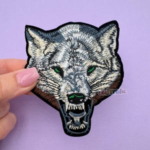 May include: Embroidered iron-on patch of a snarling wolf's head with green eyes. The wolf is white and gray with a brown muzzle. The patch is on a purple background.