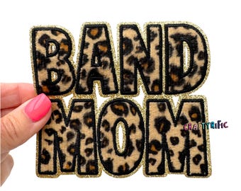 May include: A decorative patch with the words "BAND MOM" in a leopard print design. The letters are outlined in gold glitter, creating a bold and eye-catching accessory. The patch is likely for clothing or accessories.