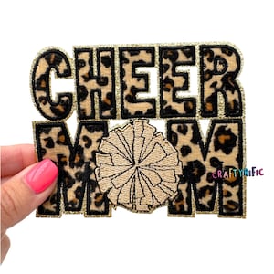 May include: A rectangular patch with the words "CHEER MOM" in a leopard print design with gold glitter trim. A tan and gold pom-pom is in the center. The patch is held by a person with pink nail polish.
