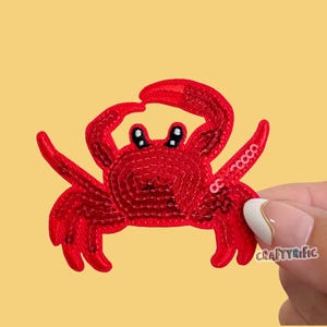 May include: A bright red crab patch featuring a sequined texture and embroidered details. The crab has two raised claws, multiple legs, and black eyes with white highlights. The text Craftyrific is visible in the lower right corner of the patch.