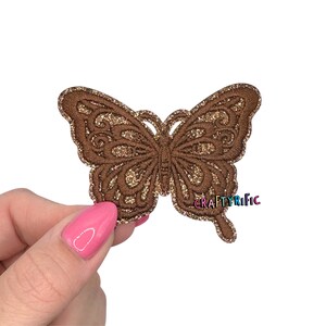 May include: A brown butterfly-shaped applique with intricate embroidered details and gold glitter accents. The applique has a detailed design and is held by a hand. The text "CRAFTYRIFIC" is visible.