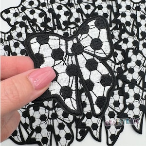 May include: A black and white checkered bow with a soccer ball pattern. The bow is made of fabric and has a stitched edge. The bow is surrounded by other soccer ball patterned fabric shapes.