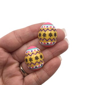 May include: A pair of pink and yellow Easter egg-shaped earrings with purple and white accents. The earrings have a zig-zag pattern and black flower designs.