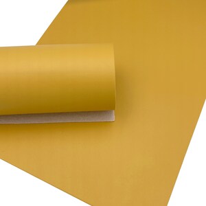 May include: Mustard yellow faux leather sheet, partially rolled, with a smooth, matte finish. The sheet is shown against a white background, highlighting the color and texture of the material. The rolled portion reveals a tan backing.