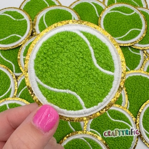 May include: A close-up of a green and white tennis ball patch with a gold glitter border. The patch is made of fuzzy fabric and has a white stitched seam. The patch is being held by a hand.