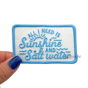 May include: Embroidered patch with the text "ALL I NEED IS Sunshine AND Salt water" in light blue script. The patch has a white background and a light blue border. The design includes a sun and waves.
