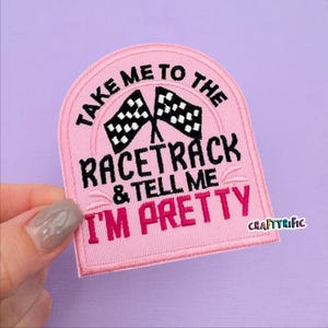 May include: Pink embroidered patch with the text "TAKE ME TO THE RACETRACK & TELL ME I'M PRETTY" and two checkered flags. The patch has a stitched border and is held by a hand against a purple background.