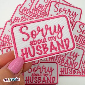 May include: White rectangular patch with pink embroidered text that reads "Sorry about my husband." The patch has a pink border and is surrounded by other similar patches. The word "CRAFTYRIFIC" is printed on the bottom left.