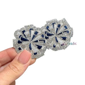 May include: Two glittery cheerleading pom-pom patches. Each patch features a silver glitter outline, white and navy blue accents, and a central design resembling a starburst. The text "CRAFTYRIFIC" is visible on the right patch.
