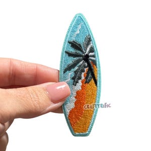 May include: Embroidered surfboard patch featuring a palm tree, waves, and a sandy beach scene. The patch has a light blue background with orange, white, and black details. The text "CraftyRific" is visible on the bottom right.