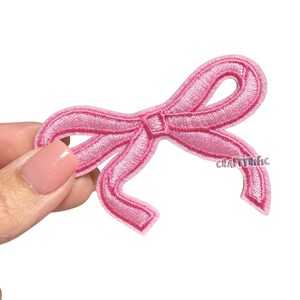 May include: A pink embroidered bow patch with the text "CRAFTYRIFIC" in the lower right corner. The bow is a light shade of pink with a detailed stitch pattern, perfect for adding to clothing or accessories.