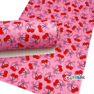 May include: Pink patterned fabric featuring red cherries with pink bows. The cherries have black stems and are scattered across the pink background. The fabric is rolled up, showing the underside. The text "CRAFTYRIFIC" is visible.